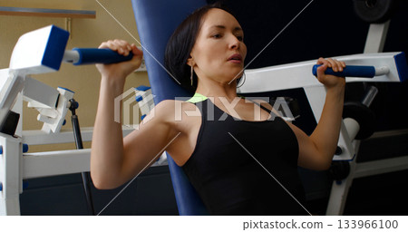 Caucasian woman in excellent physical shape during training in the gym. Deltoid press. Physical education as a norm of life to maintain health. Close-up photo. 133966100