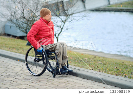 Man in wheelchair enjoys calm day by the water while wearing red jacket and brown hat Man in wheelchair enjoys calm day by the water while wearing red jacket and brown hat 133968139