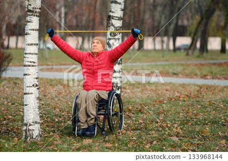 Senior person in wheelchair exercises outdoors using resistance band on a fall day Senior person in wheelchair exercises outdoors using resistance band on a fall day 133968144