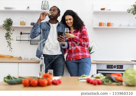 A couple is in their kitchen, looking at a phone while enjoying wine. Fresh vegetables are on the counter. A couple is in their kitchen, looking at a phone while enjoying wine. Fresh vegetables are on the counter. 133968310