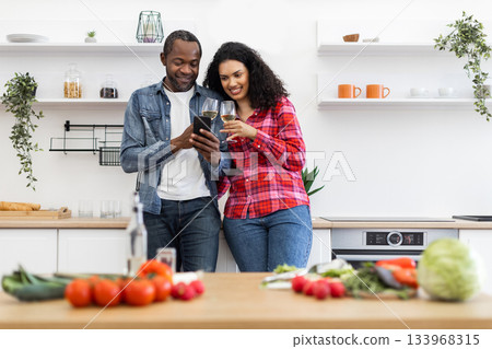 A diverse couple enjoys wine and looks at a phone in a modern kitchen with fresh produce. 133968315