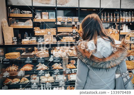 A woman stands in a bakery examining a variety of sweet cakes, croissants, and buns. She is deciding on a dessert to purchase for dinner 133970489