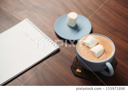 Hot chocolate in a blue cup with marshmallow and notepad on walnut table 133971898