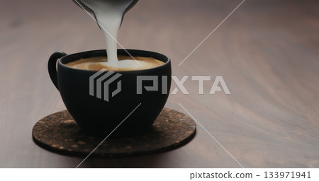 pour steamed milk into cappuccino black cup on walnut table pour steamed milk into cappuccino black cup on walnut table 133971941