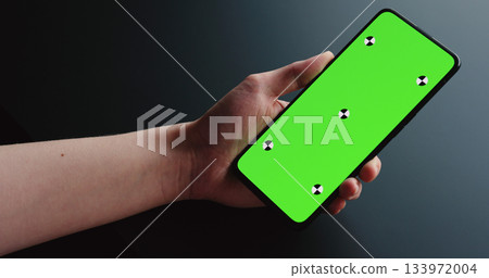 young woman sitting at the black table near window and holding smartphone with vertical green screen 133972004