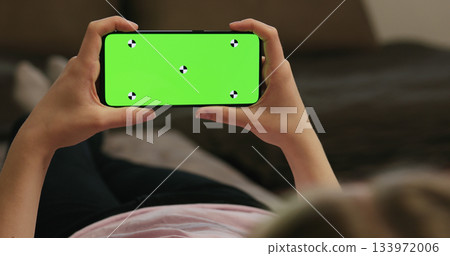 young woman lying on a couch and holding smartphone with horizontal green screen 133972006