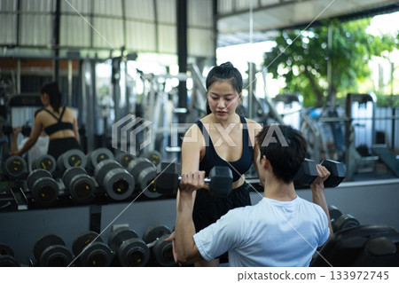 A woman assists and checks a man's dumbbell lifting form during a workout at the gym A woman assists and checks a man's dumbbell lifting form during a workout at the gym 133972745