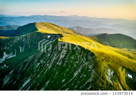Aerial view of rugged mountain range with lush green slopes at sunset. Landscape showcases beauty of untouched nature with distant peaks and valleys bathed in soft sunlight. Carpathians, Chornogora. 133973014
