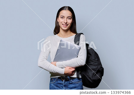 Portrait of beautiful female teenage student with backpack, laptop on gray background Portrait of beautiful female teenage student with backpack, laptop on gray background 133974366