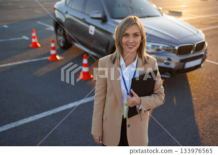 Driving instructor standing in front of learner vehicle during practice session Driving instructor standing in front of learner vehicle during practice session 133976565