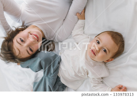 Happy mother and baby lying together on a soft white bed, enjoying a warm and cozy family moment filled with love, comfort, connection, and natural light. 133978313