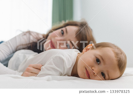 Cozy moment of a young woman and baby relaxing together at home, lying on a soft bed and smiling in natural light. Cozy moment of a young woman and baby relaxing together at home, lying on a soft bed and smiling in natural light. 133978325