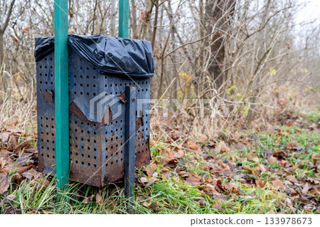 A trash bin stands in a woodland area with rust and a black bag on top. Fall leaves cover the ground, and the sky looks gray A trash bin stands in a woodland area with rust and a black bag on top. Fall leaves cover the ground, and the sky looks gray 133978673
