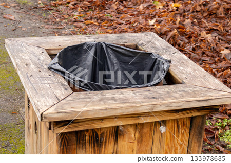 A wooden trash can with a black bag sits in a park area with fallen leaves around it. The scene shows late autumn conditions 133978685