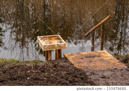 A wooden platform and simple stairs lead to water. Trees reflect in the water. The ground is moist and earthy A wooden platform and simple stairs lead to water. Trees reflect in the water. The ground is moist and earthy 133978688