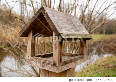 A birdhouse is placed on a post near a pond surrounded by trees and grass. The scene shows nature in a quiet moment A birdhouse is placed on a post near a pond surrounded by trees and grass. The scene shows nature in a quiet moment 133978690