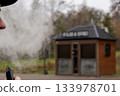 A person smokes an electronic device and exhales vapour outside a small wooden building in a park. The scene takes place in daylight 133978701