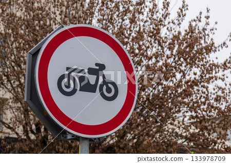 A round traffic sign shows a motorcycle symbol with a red circle around it, placed near bare trees on a cloudy day A round traffic sign shows a motorcycle symbol with a red circle around it, placed near bare trees on a cloudy day 133978709