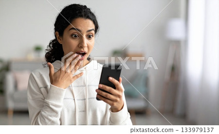 A young woman stands indoors holding a smartphone, her eyes wide, mouth open in shock, Warm ambient lighting and a softly blurred modern interior create a lively backdrop for her expressive reaction A young woman stands indoors holding a smartphone, her eyes wide, mouth open in shock, Warm ambient lighting and a softly blurred modern interior create a lively backdrop for her expressive reaction 133979137