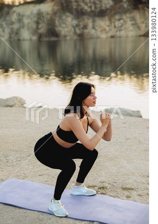 Woman performing squats by the serene lake at sunset, showcasing fitness and dedication to a healthy lifestyle in nature 133980124