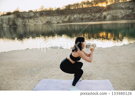 Woman exercising with weights by the lake during sunset near cliffs in a tranquil setting Woman exercising with weights by the lake during sunset near cliffs in a tranquil setting 133980126