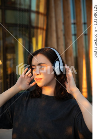 Young woman wearing headphones enjoys music outdoors during golden hour near modern architecture 133980128