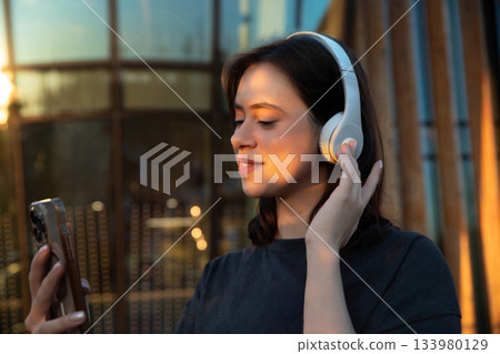 Young woman listens to music with headphones while using a smartphone in a bright urban setting during sunset Young woman listens to music with headphones while using a smartphone in a bright urban setting during sunset 133980129