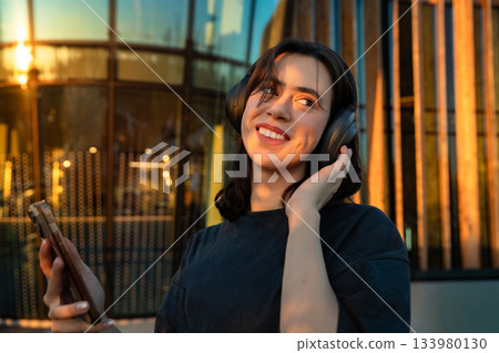 Young woman enjoys music while listening on headphones at sunset near a modern building in a vibrant urban setting Young woman enjoys music while listening on headphones at sunset near a modern building in a vibrant urban setting 133980130