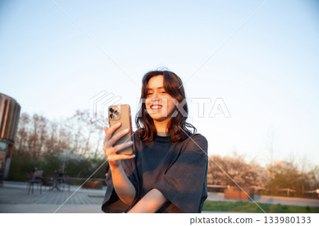 Young woman enjoying a sunset while using her smartphone in a peaceful outdoor setting with blooming trees and clear sky 133980133
