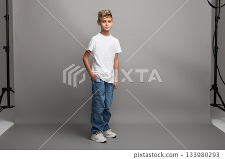 Young boy model with stylish haircut standing in studio. Kid modeling new clothing or showcasing child fashion with copy space on gray background Young boy model with stylish haircut standing in studio. Kid modeling new clothing or showcasing child fashion with copy space on gray background 133980293