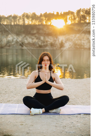 Woman practicing yoga by a tranquil lake at sunset, reflecting calmness and connection with nature during golden hour 133980756