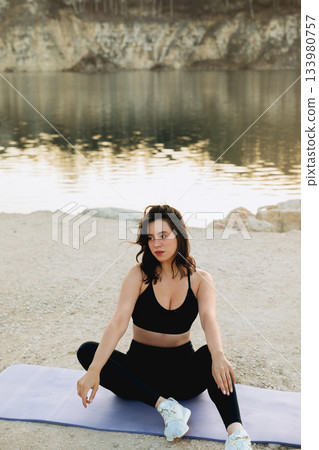 Woman practicing yoga by the calm lake at sunrise, surrounded by nature in a peaceful outdoor setting with rocky cliffs Woman practicing yoga by the calm lake at sunrise, surrounded by nature in a peaceful outdoor setting with rocky cliffs 133980757