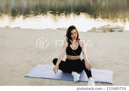 Woman practicing yoga by a calm lake during the late afternoon, focusing on relaxation and mindfulness 133980758