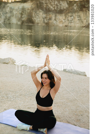 Woman practicing yoga at sunset by a serene lake in a rocky outdoor setting, embracing tranquility and mindfulness during her session Woman practicing yoga at sunset by a serene lake in a rocky outdoor setting, embracing tranquility and mindfulness during her session 133981020