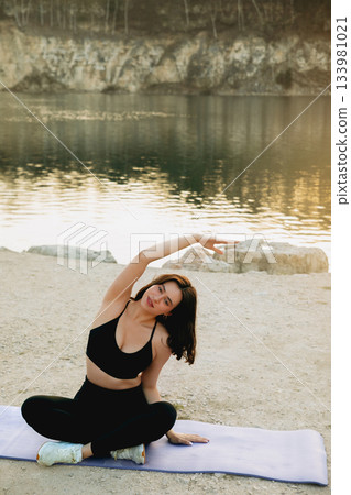Woman practicing yoga by the lake during sunset, showcasing flexibility and tranquility in a serene natural setting Woman practicing yoga by the lake during sunset, showcasing flexibility and tranquility in a serene natural setting 133981021