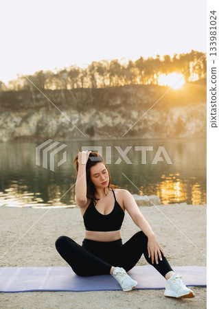 Practicing yoga at sunrise by the lake with warm sunlight reflecting on the water and trees in the background Practicing yoga at sunrise by the lake with warm sunlight reflecting on the water and trees in the background 133981024