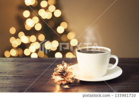 White cup of hot coffee with pine cone on wooden table and Christmas tree bokeh lights behind. Warm cozy winter holiday background. 133981710