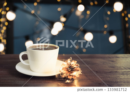 White cup of hot coffee on wooden table with pine cone and warm festive lights. Cozy winter atmosphere, christmas mood, holiday background. White cup of hot coffee on wooden table with pine cone and warm festive lights. Cozy winter atmosphere, christmas mood, holiday background. 133981712