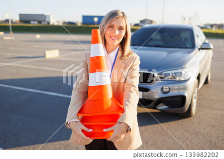 Driving instructor holding safety cones near training vehicle in outdoor parking area Driving instructor holding safety cones near training vehicle in outdoor parking area 133982202