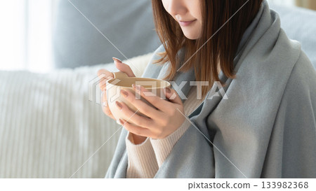 A woman wearing a blanket and drinking a hot drink 133982368