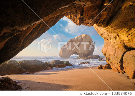 At Praia da Santa Cruz, Penedo do Guincho stands majestically as waves crash into the sandy beach beneath a colorful sunset. 133984076