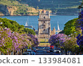 Scenic view of Belem Tower surrounded by blooming jacaranda trees, with sailboats on the Tagus River during sunset in Lisbon, Portugal. 133984081