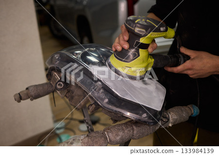 Hands refining car lamps professionally, Orbital buffer restoring clarity to headlights, Closeup of technician smoothing car headlight with orbital buffer and polishing tools Hands refining car lamps professionally, Orbital buffer restoring clarity to headlights, Closeup of technician smoothing car headlight with orbital buffer and polishing tools 133984139