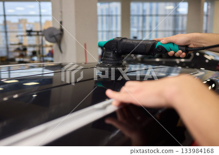Polishing black car surface thoroughly, Detailed buffing to achieve showroom quality shine, Expert technician uses orbital buffer and foam pad to enhance vehicle gloss 133984168
