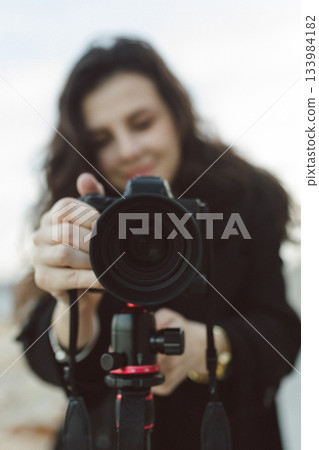 Female photographer adjusting professional camera on tripod outdoors by the sea, focusing the lens while preparing for a creative photo shoot on a rocky coastline. 133984182