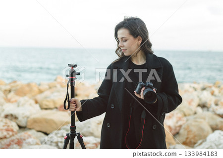 Female photographer adjusting professional camera on tripod outdoors by the sea, focusing the lens while preparing for a creative photo shoot on a rocky coastline. 133984183