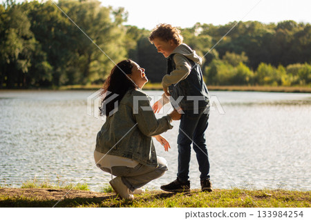 Curious boy points across lake while his mother kneels beside him, sharing moment of discovery. Perfect for motherhood bonding themes. High quality photo. Curious boy points across lake while his mother kneels beside him, sharing moment of discovery. Perfect for motherhood bonding themes. High quality photo. 133984254