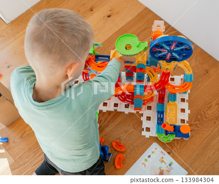 A young child plays with a colorful construction toy set on a wooden floor. Bright pieces and an instruction sheet highlight creativity, motor skill development and early learning in a playful home 133984304