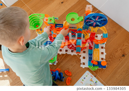 A toddler reaches for pieces of a colorful construction set laid out on a wooden floor. The scene captures focused play, early problem-solving and joyful learning in a bright home environment. A toddler reaches for pieces of a colorful construction set laid out on a wooden floor. The scene captures focused play, early problem-solving and joyful learning in a bright home environment. 133984305