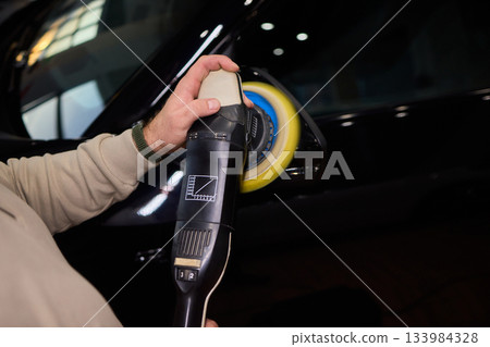 Polishing black car surface thoroughly, Detailed buffing to achieve showroom quality shine, Expert technician uses orbital buffer and foam pad to enhance vehicle gloss 133984328